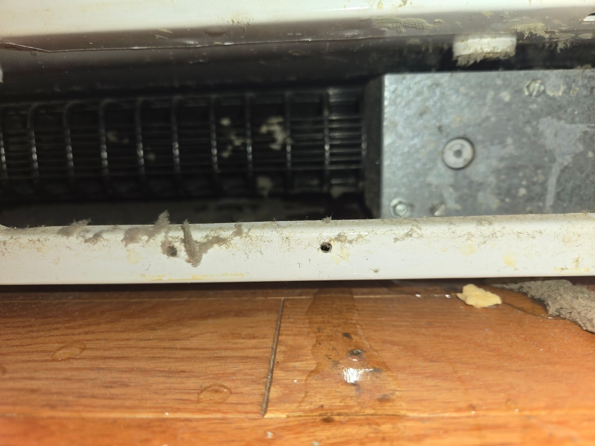 Damaged metal vent or appliance opening above a wooden floor, with rust and dirt visible.