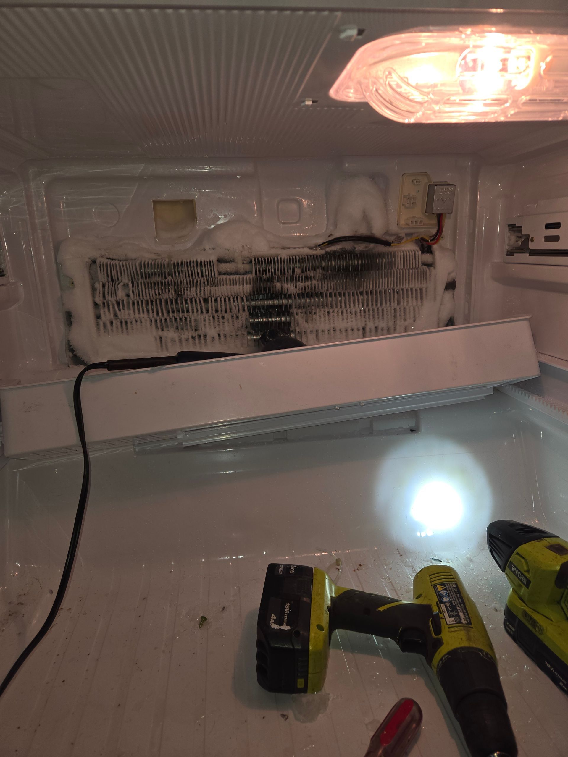 Inside a freezer: frosted evaporator coils, a light bulb, and a cordless drill on the shelf.