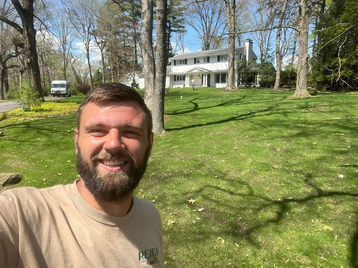 Man smiling in front of a white house on a sunny day; green lawn, trees, and blue sky.