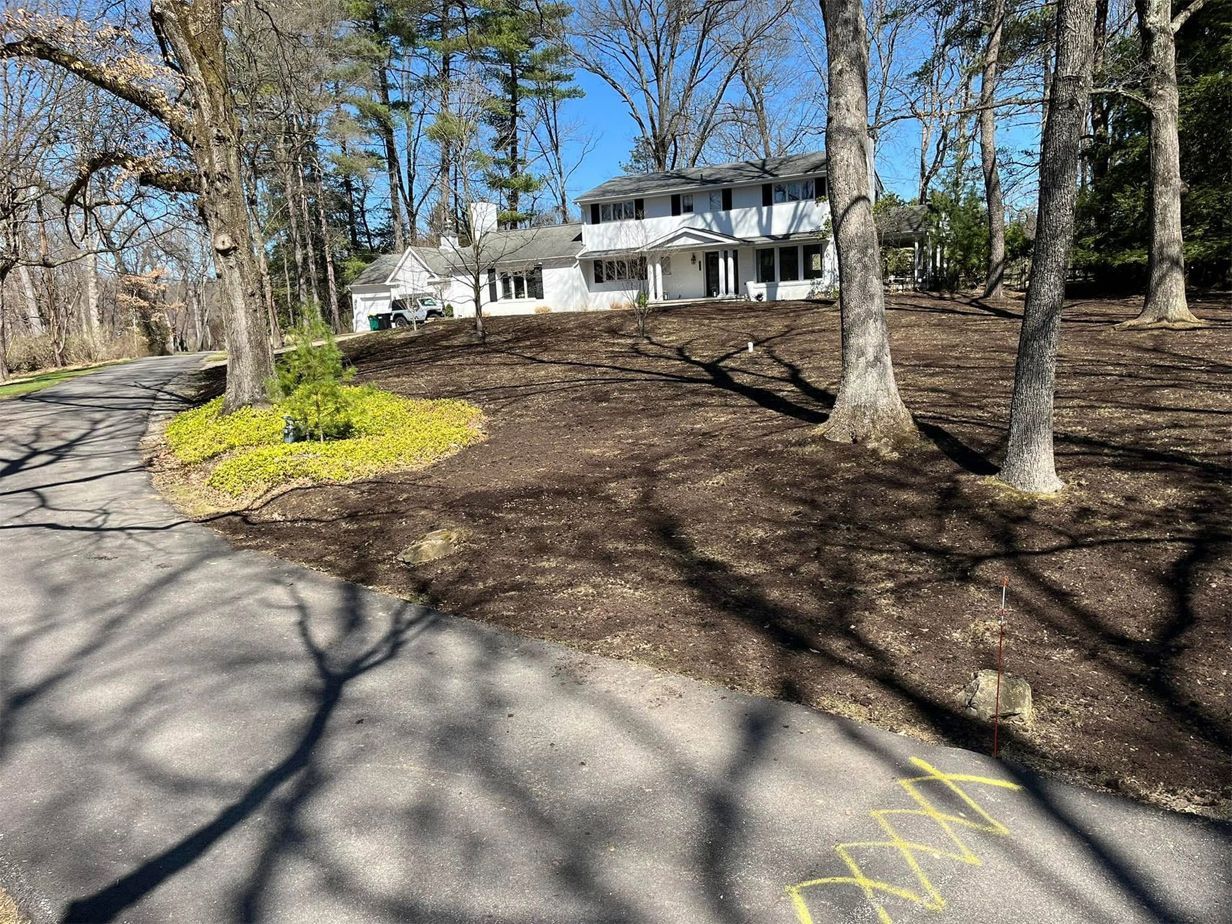 White house on a hill, surrounded by trees and newly mulched landscaping, viewed from a road.