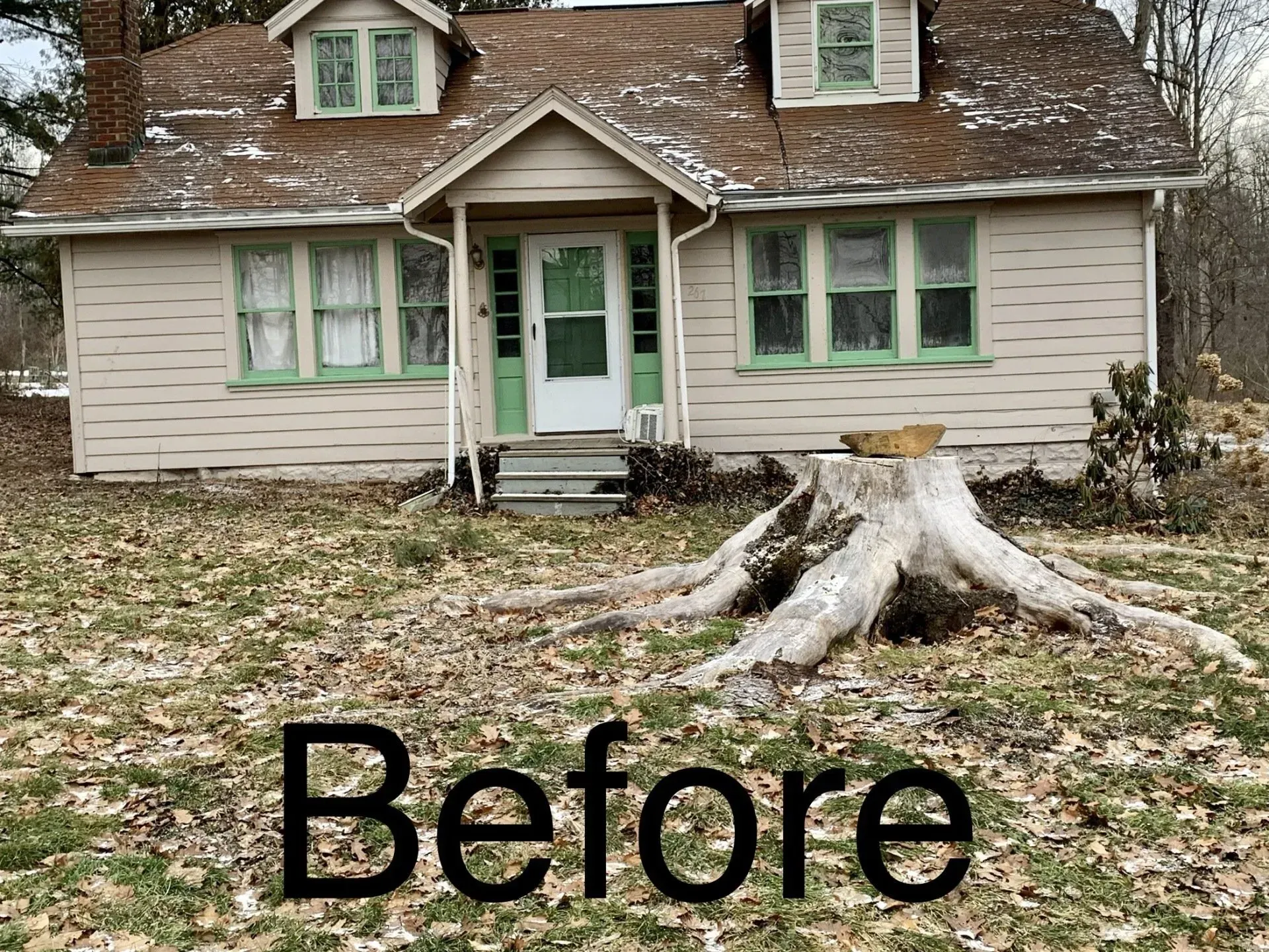 A weathered cottage with green trim and a large tree stump in the front yard.
