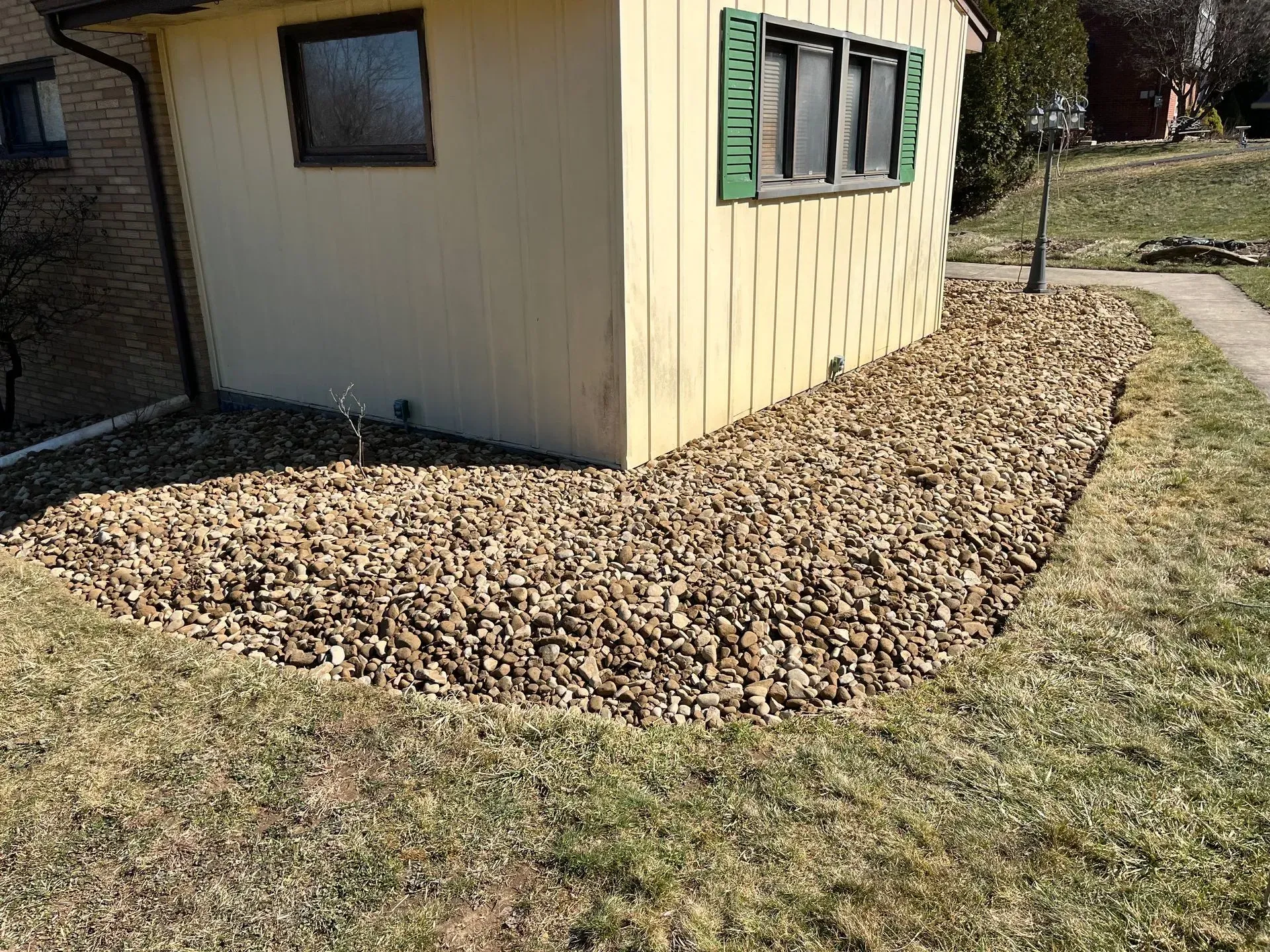 Building corner with rock bed bordering grass. Beige siding, brown rocks, and green shutters.