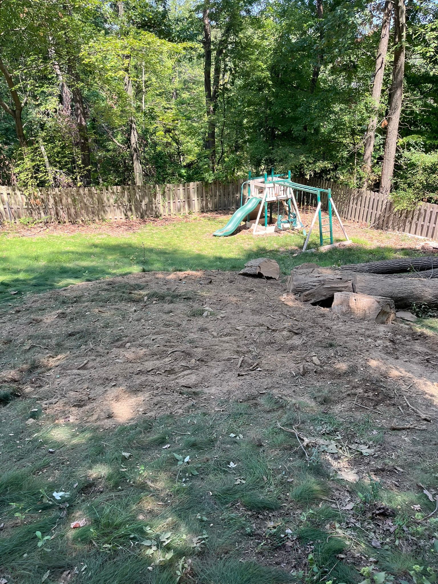 A backyard with a dirt pile, a play set, and a wooden fence surrounded by trees.