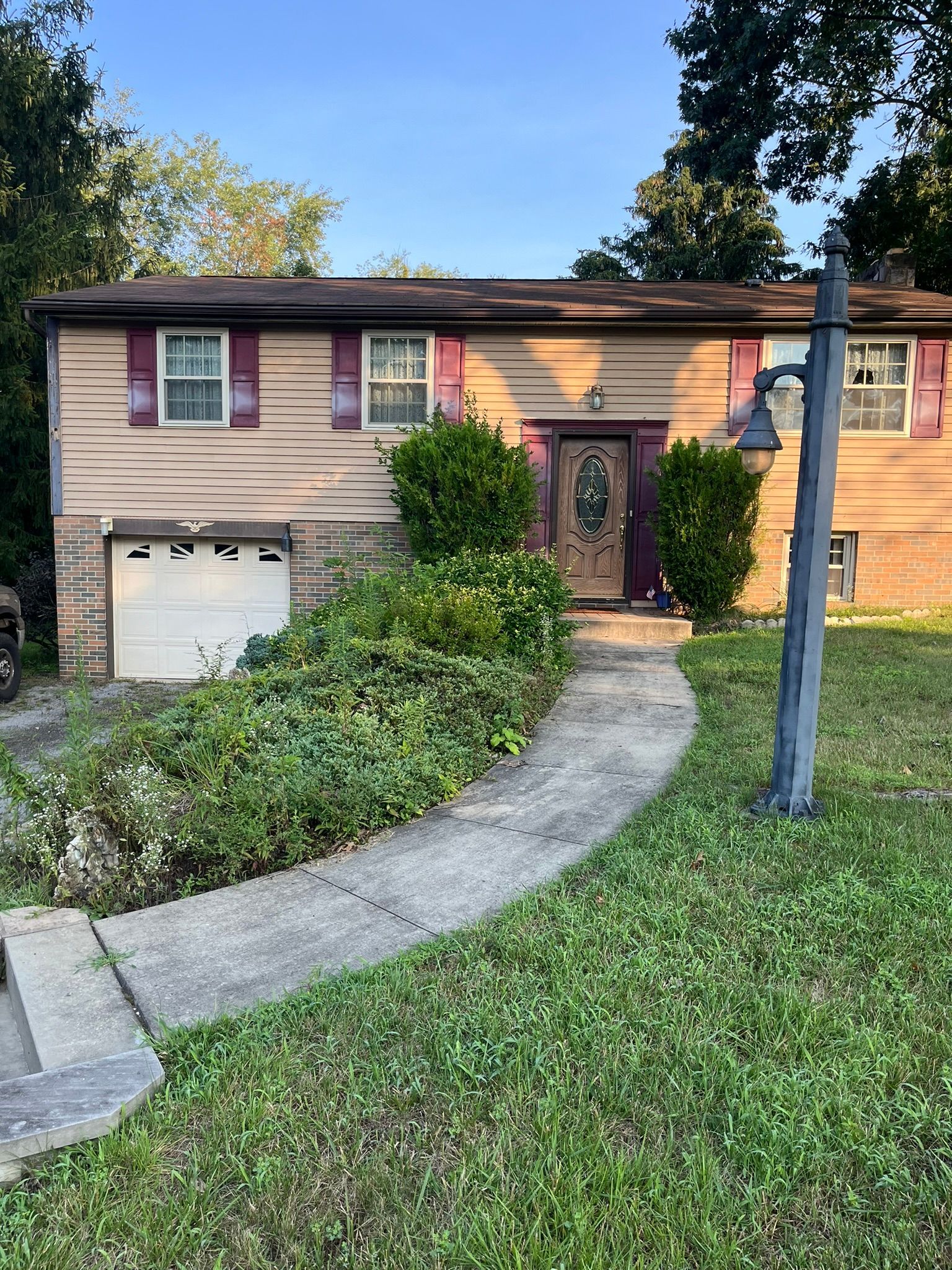 Tan house with red shutters, overgrown plants, and a curved walkway.