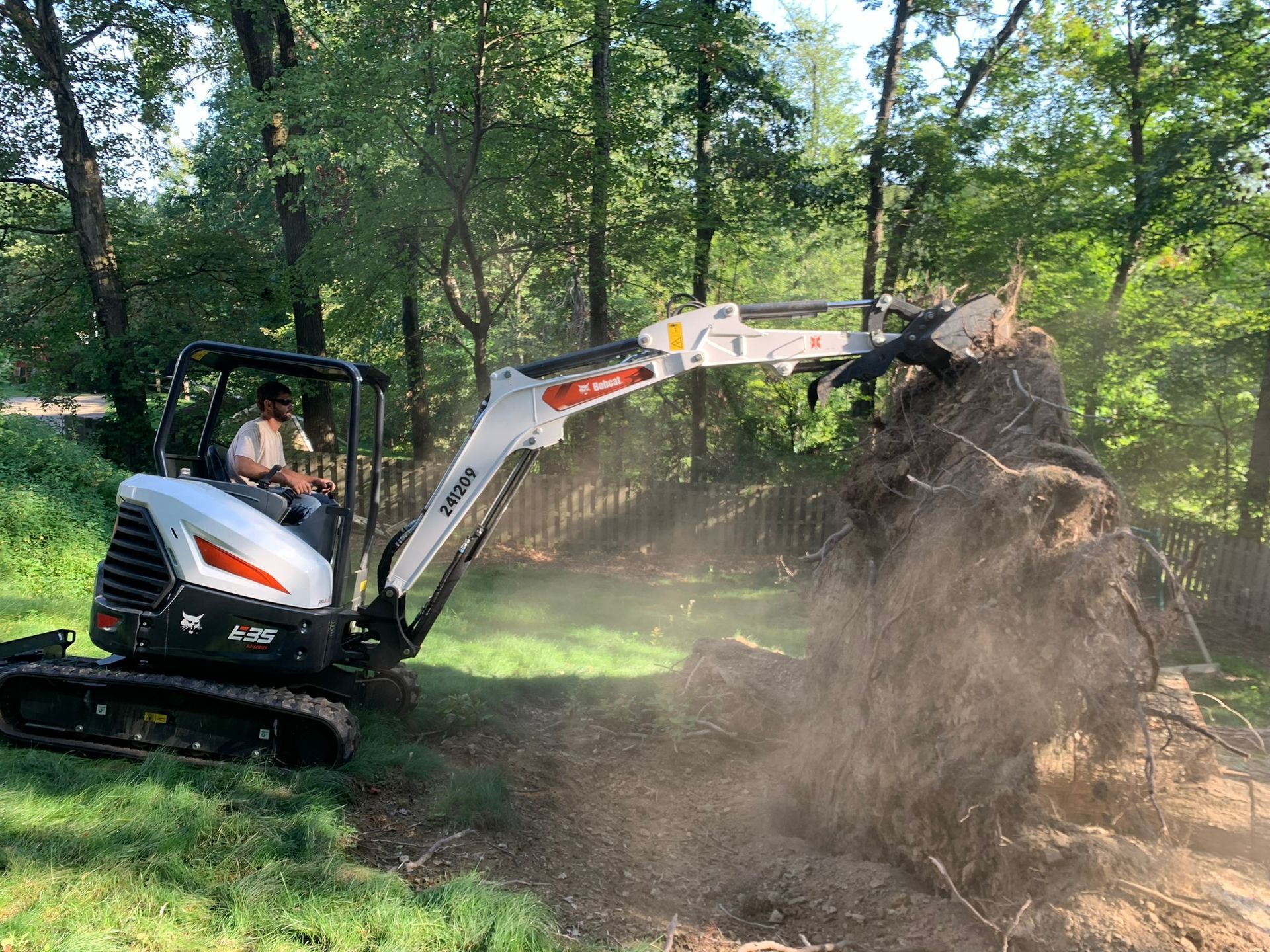 A Bobcat excavator uproots a tree stump in a grassy area, kicking up dirt. A person operates the machine.