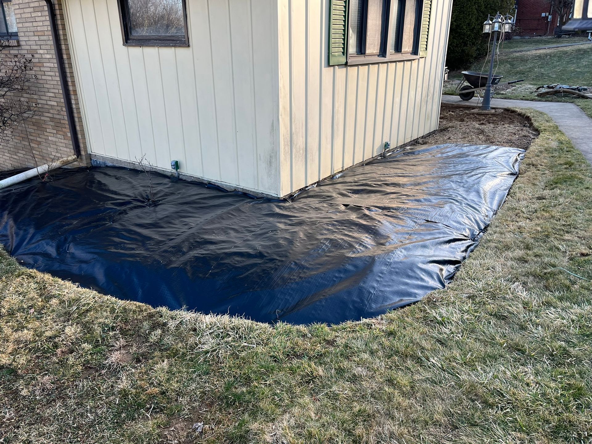 Black tarp covering a flower bed next to a light yellow house with green trim.