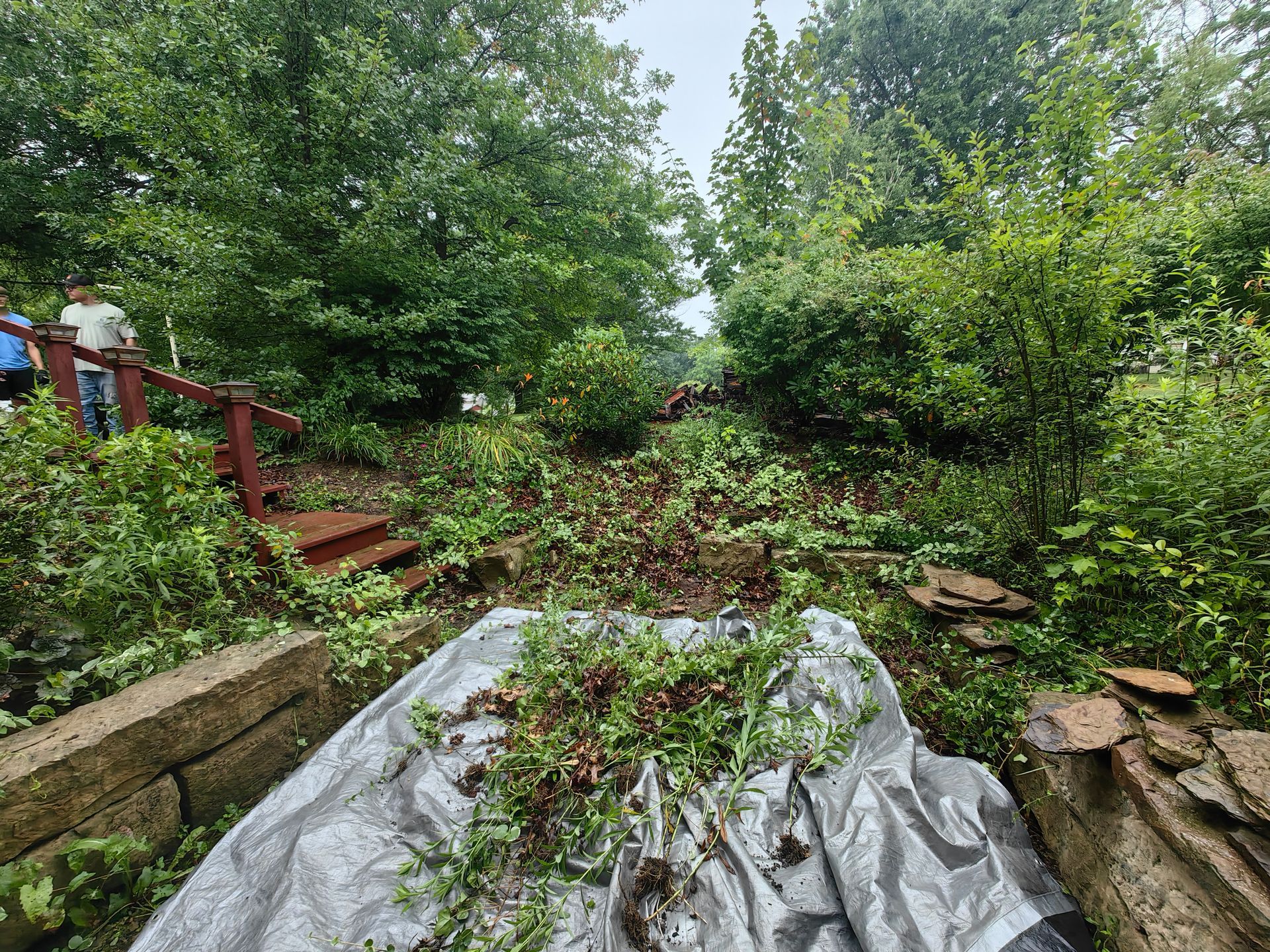 Debris covers a silver tarp on a hill, with greenery, a red staircase, and people in a wooded area.