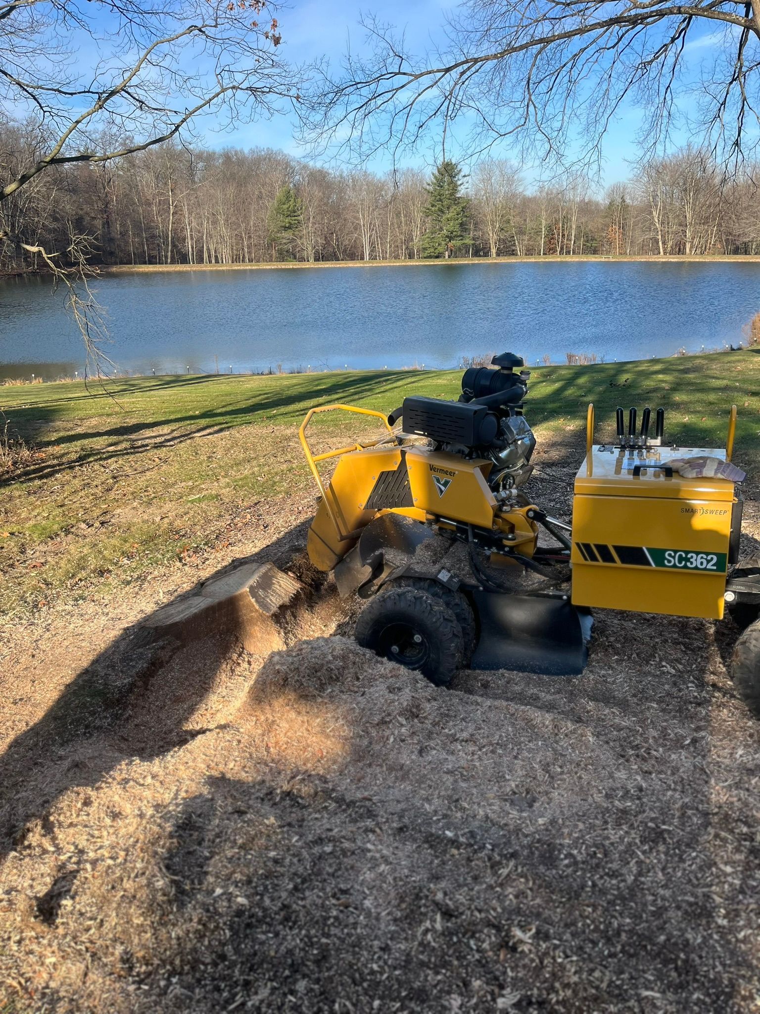 A yellow stump grinder at work next to a lake, creating wood chips.