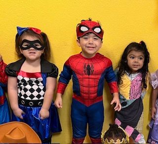 Three children in costumes: racer, Spider-Man, and Sally from 'Nightmare Before Christmas' posing against a yellow wall.
