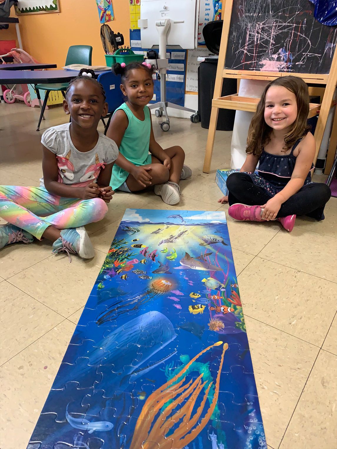 Three children sitting by a long ocean-themed puzzle on a floor; smiles.