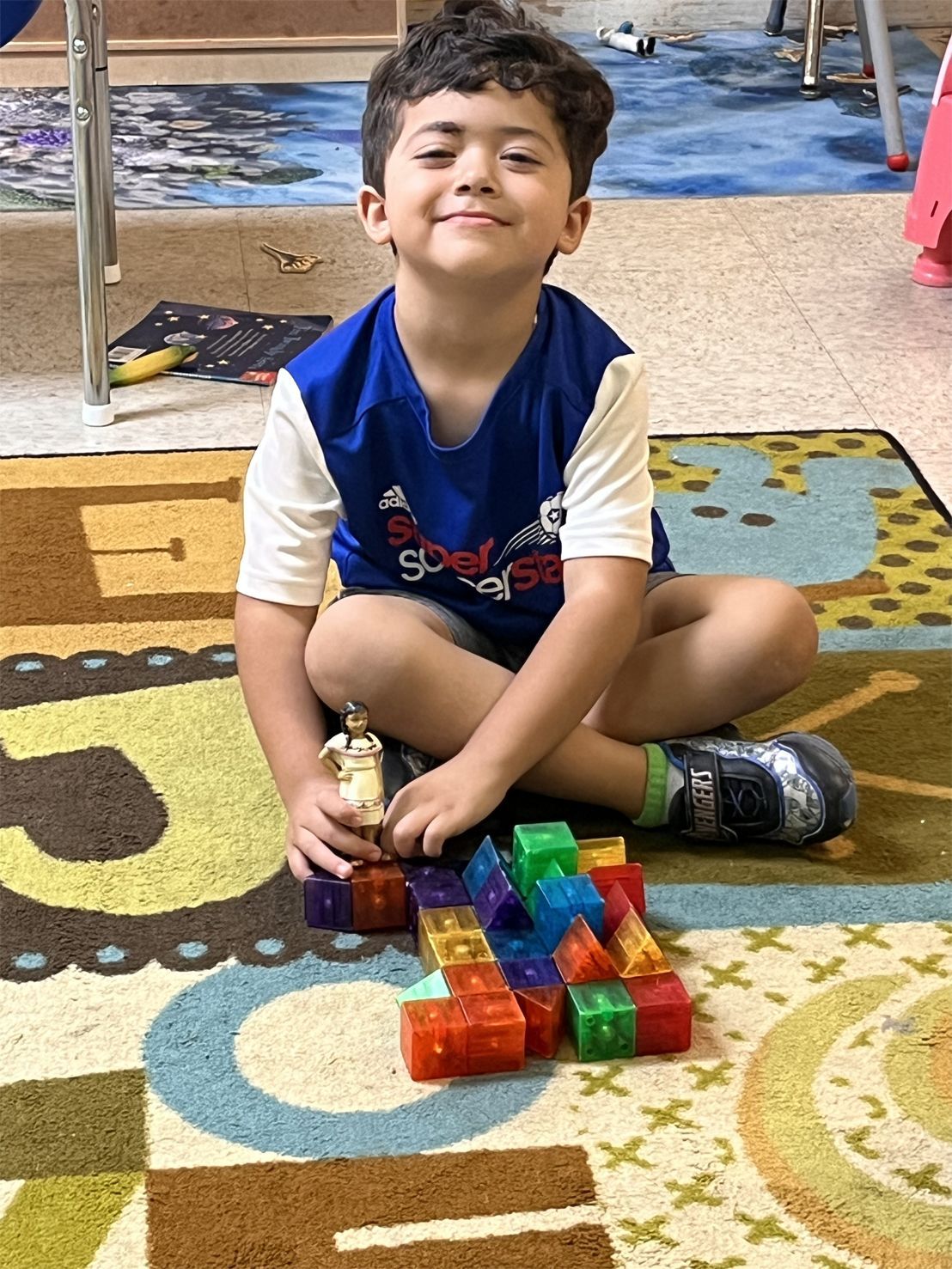 Boy sitting cross-legged smiles at the camera, building with colorful blocks on a patterned rug.