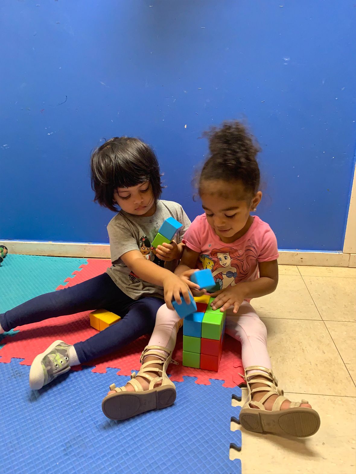 Two children building with colorful blocks on a blue and yellow mat. One child has dark hair, the other's is in a bun.