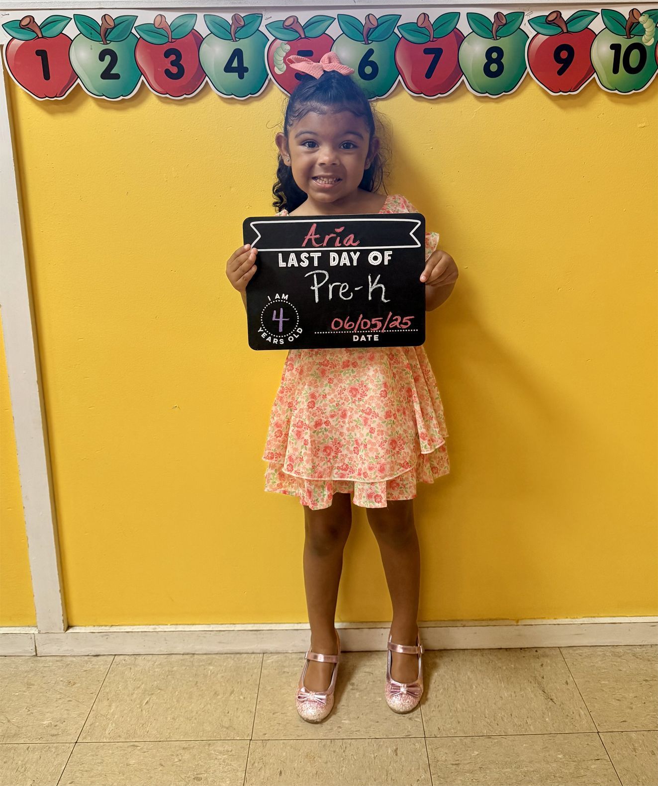 Girl holding a sign, wearing dress and shoes, standing against yellow wall with number banner.