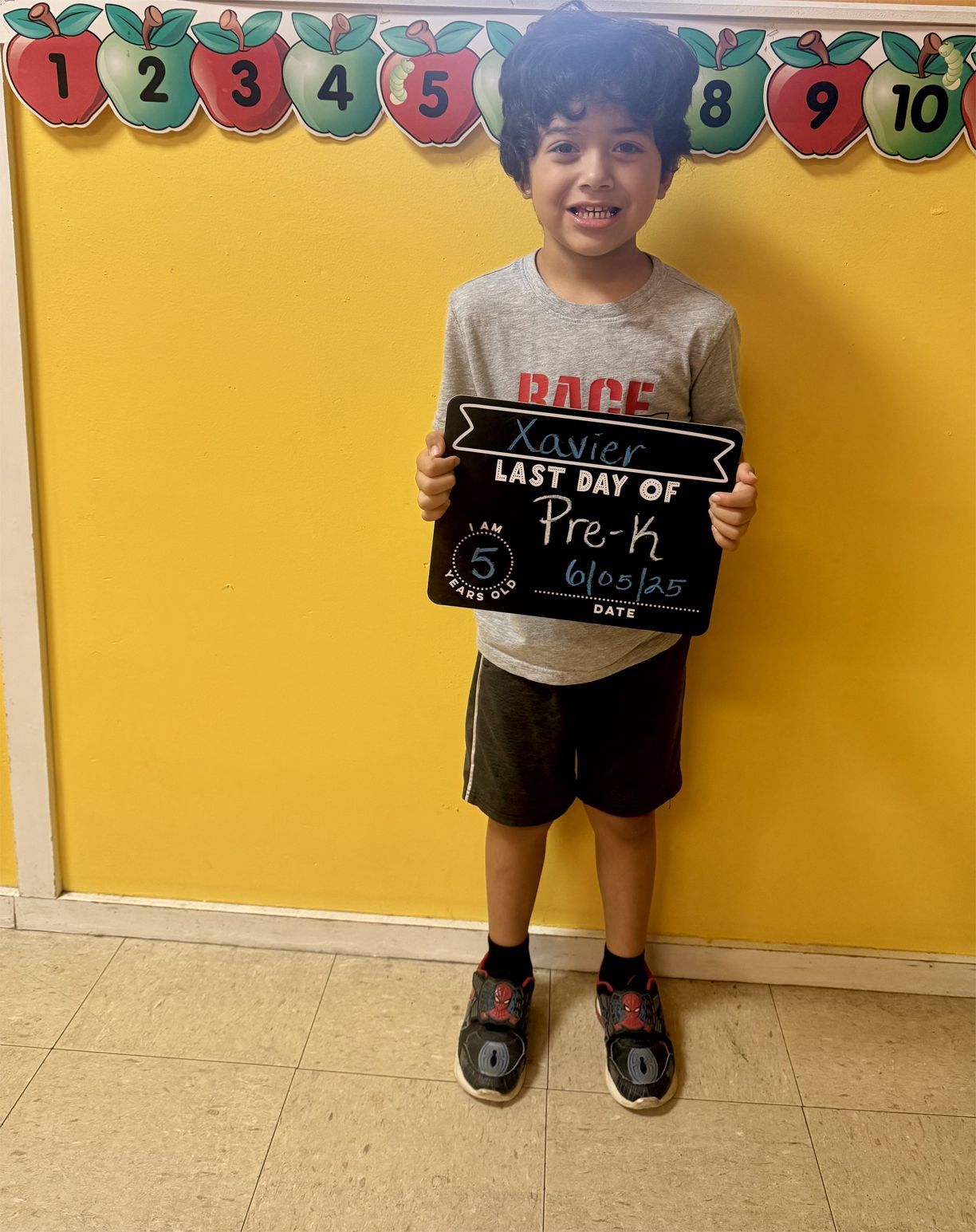 Young boy holding a chalkboard sign for his last day of Pre-K, smiling in front of a yellow wall.