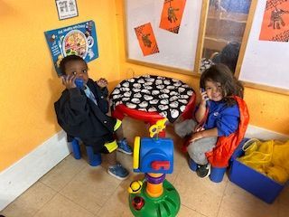 Two children sit at a table, holding toy phones. A toy mailbox is in the foreground. Brightly colored room.
