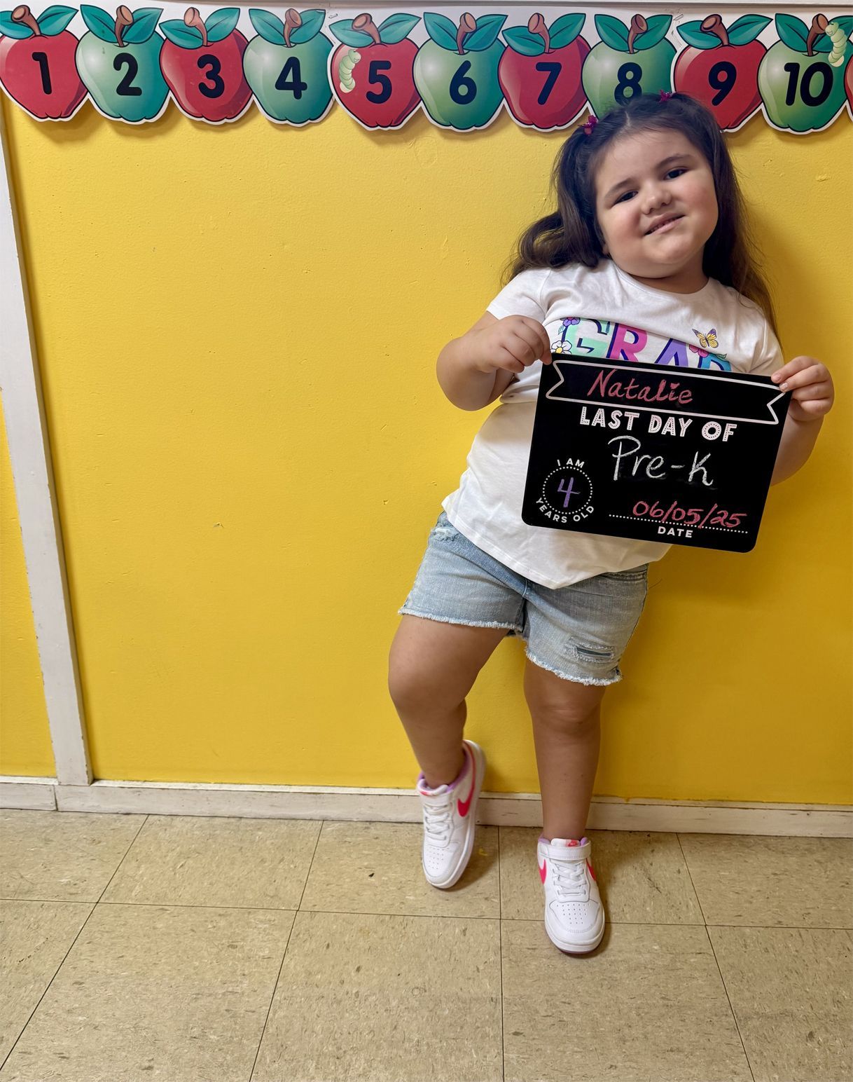A girl holding a sign stands in front of a yellow wall with apple cutouts.