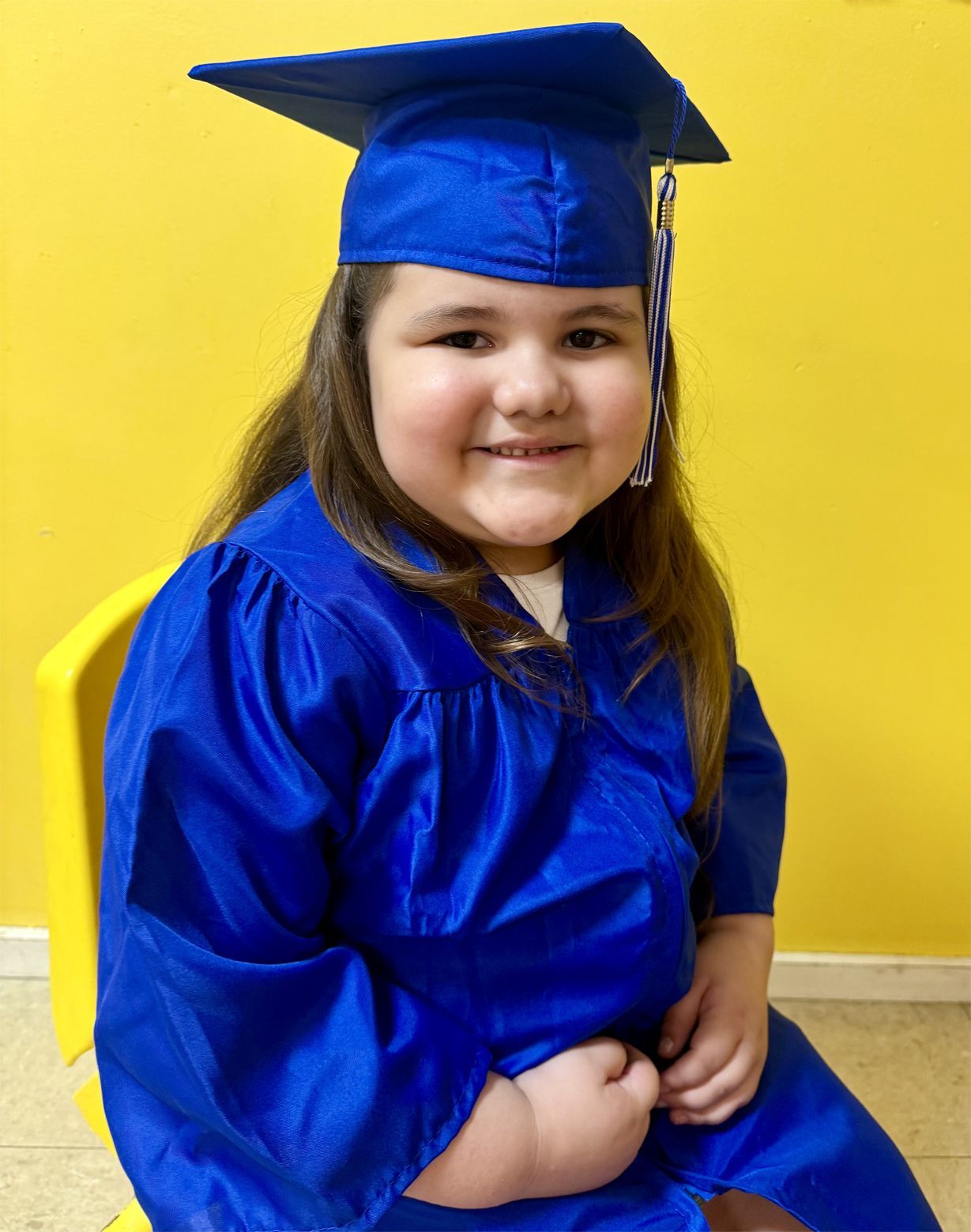 Child in a blue graduation cap and gown smiles, sitting in a yellow-walled room.