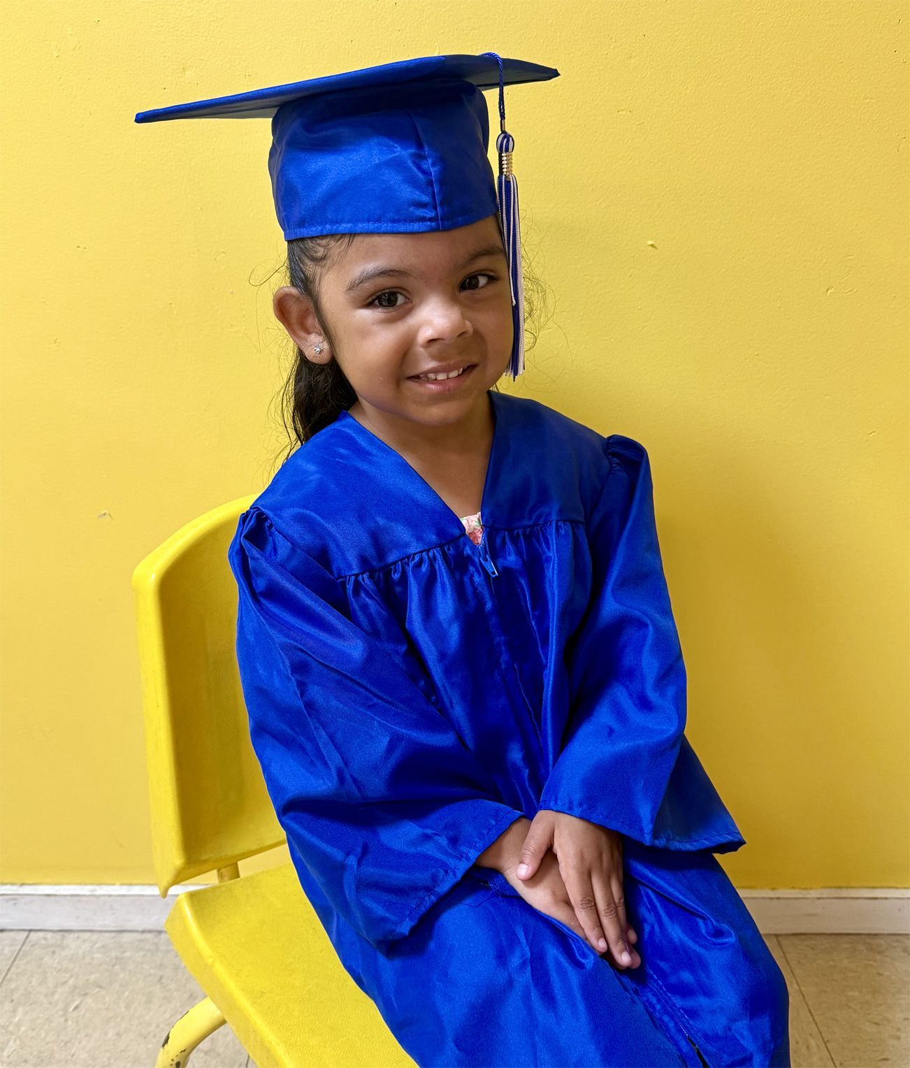 Young child in a blue graduation cap and gown smiles while seated on a yellow chair. Yellow background.