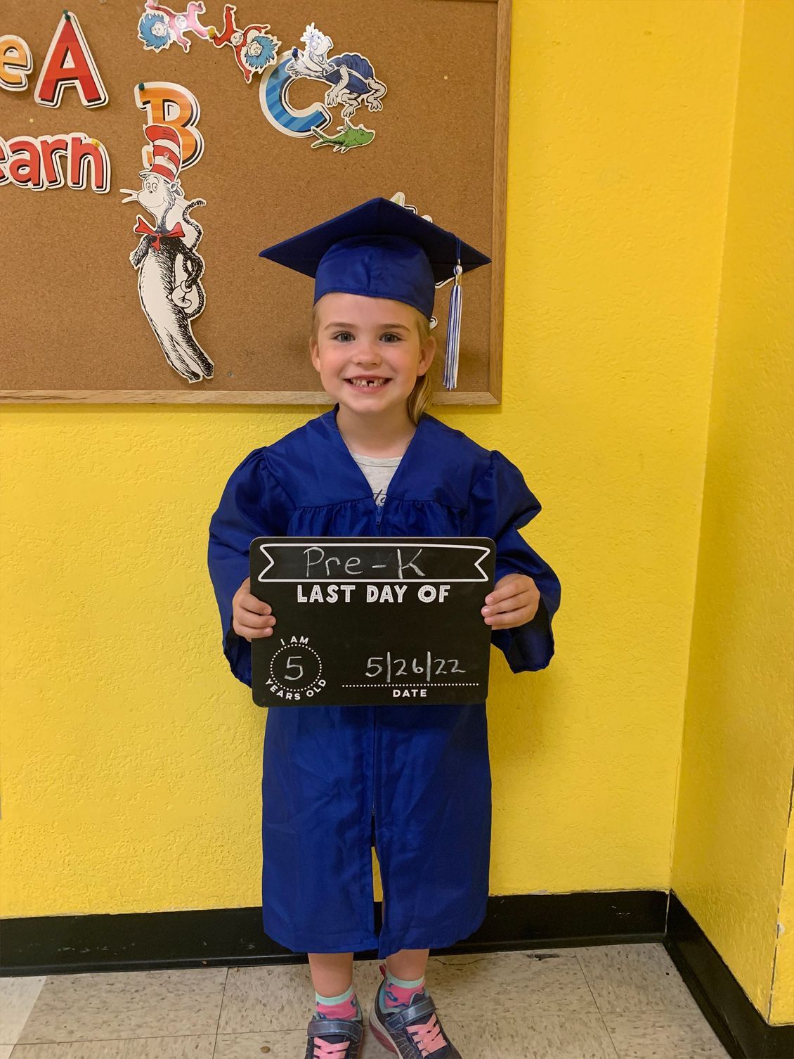 Child in blue graduation cap and gown holding a sign, smiling in front of a yellow wall.