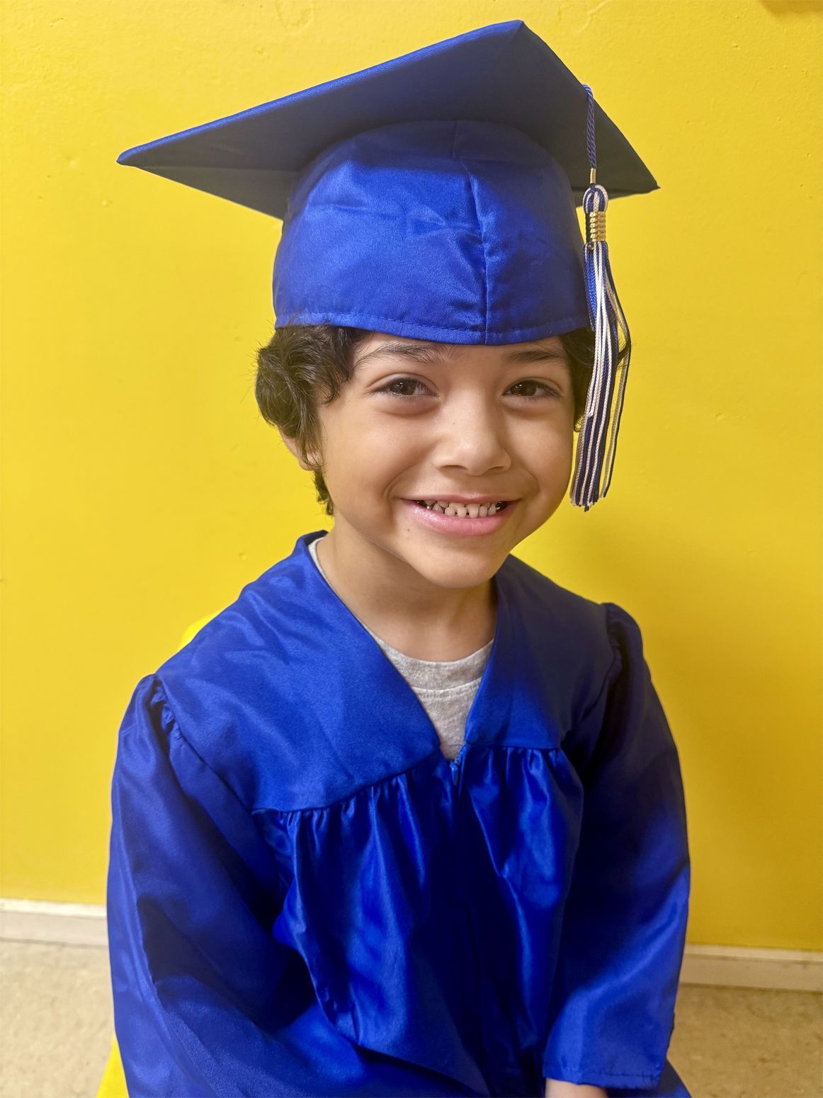Child in blue graduation cap and gown, smiling in front of a yellow background.