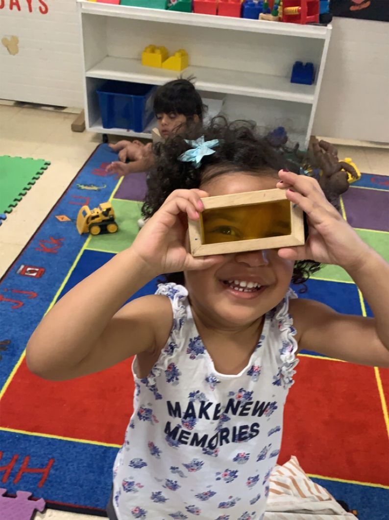 Smiling child holding a rectangular object up to their eyes, indoors with colorful mats and toys.