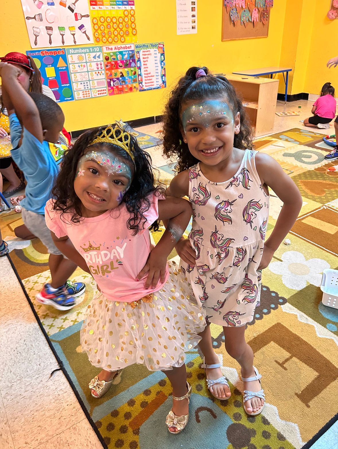 Two girls with glitter face paint smile, posing indoors on a patterned rug.