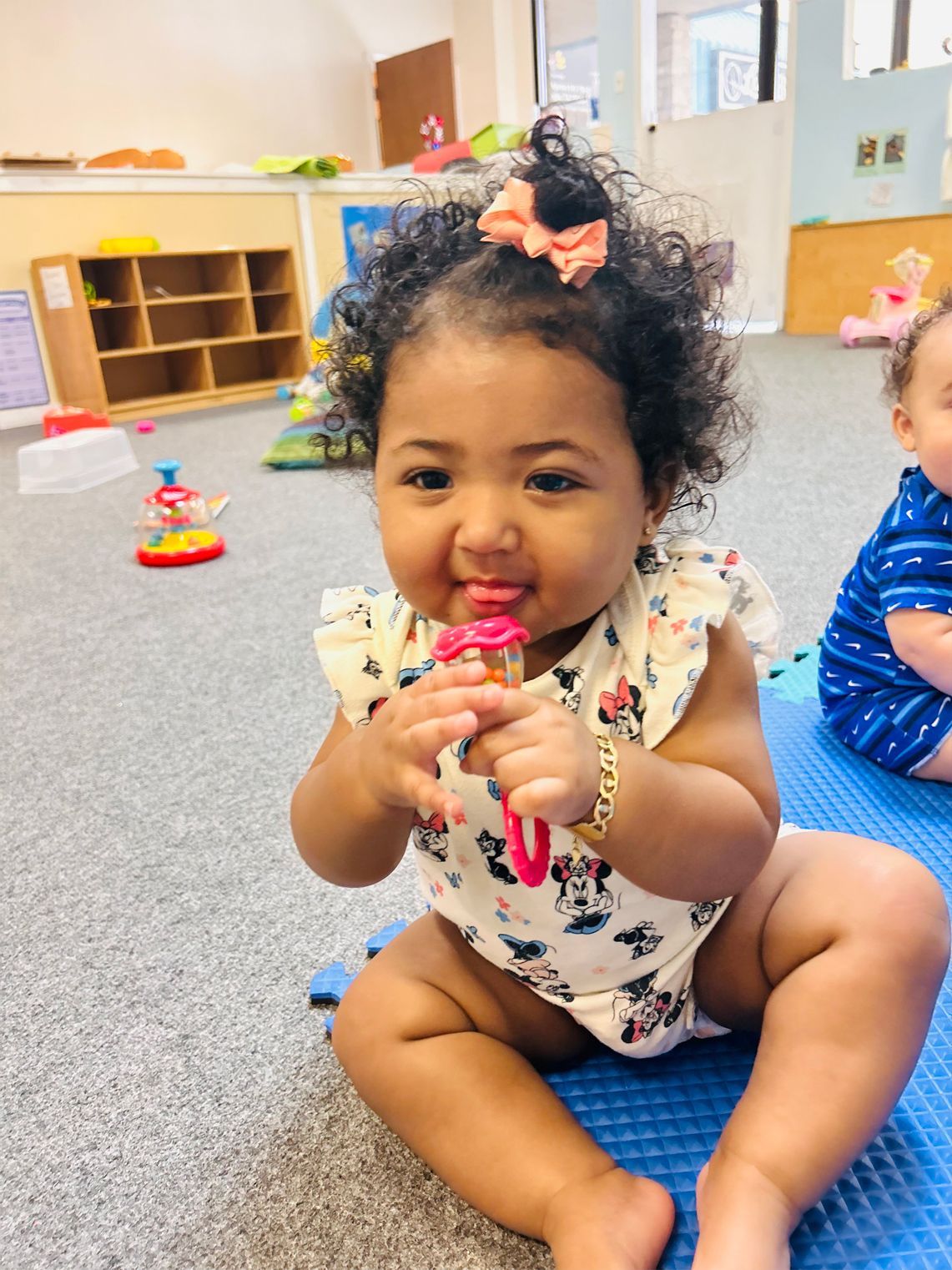 Smiling baby holding toy, sitting on blue mat in a playroom, wearing a light-colored outfit with a pink bow.