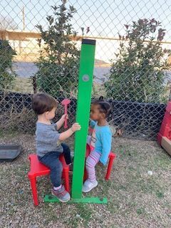 Two children play with a green pole, sitting on red chairs outdoors.