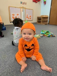 Baby in a pumpkin costume sits on a gray floor, smiling. Another child is in the background.