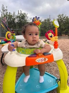 A baby sits in a colorful activity center outdoors, looking forward with a serious expression.