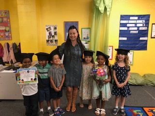 A teacher and six children in graduation caps pose indoors in front of a yellow wall.