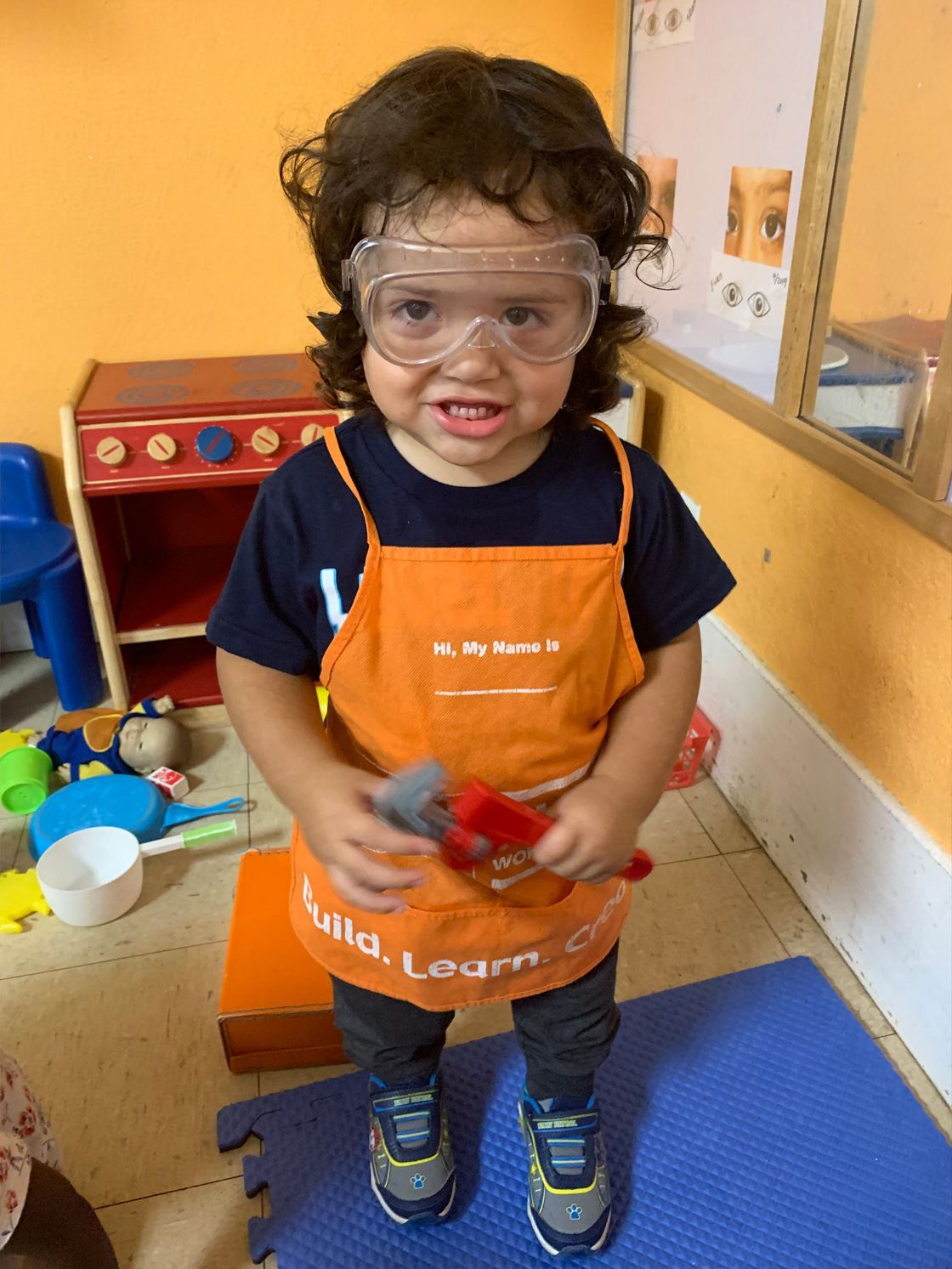 Child in orange apron and safety goggles, holding wrench, smiling, playroom setting.