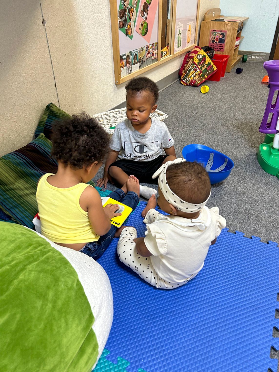 Three toddlers sitting on a blue mat, looking at a book and toys in a playroom.