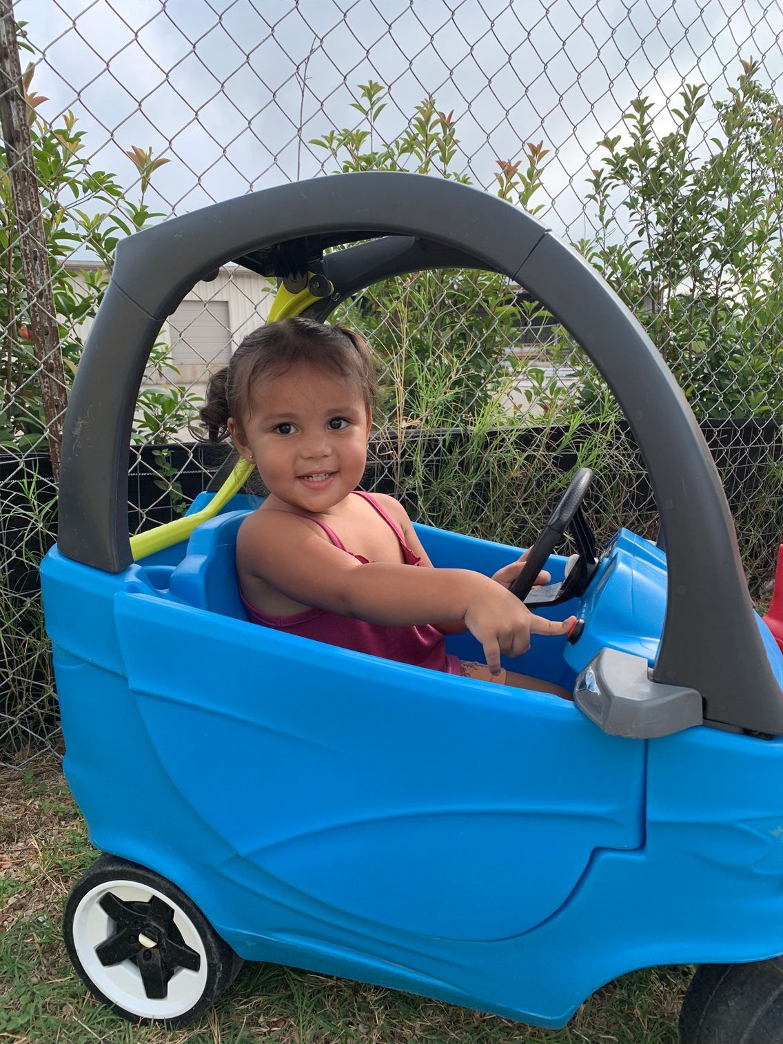 Young child smiling and pointing while sitting in a blue toy car outdoors.