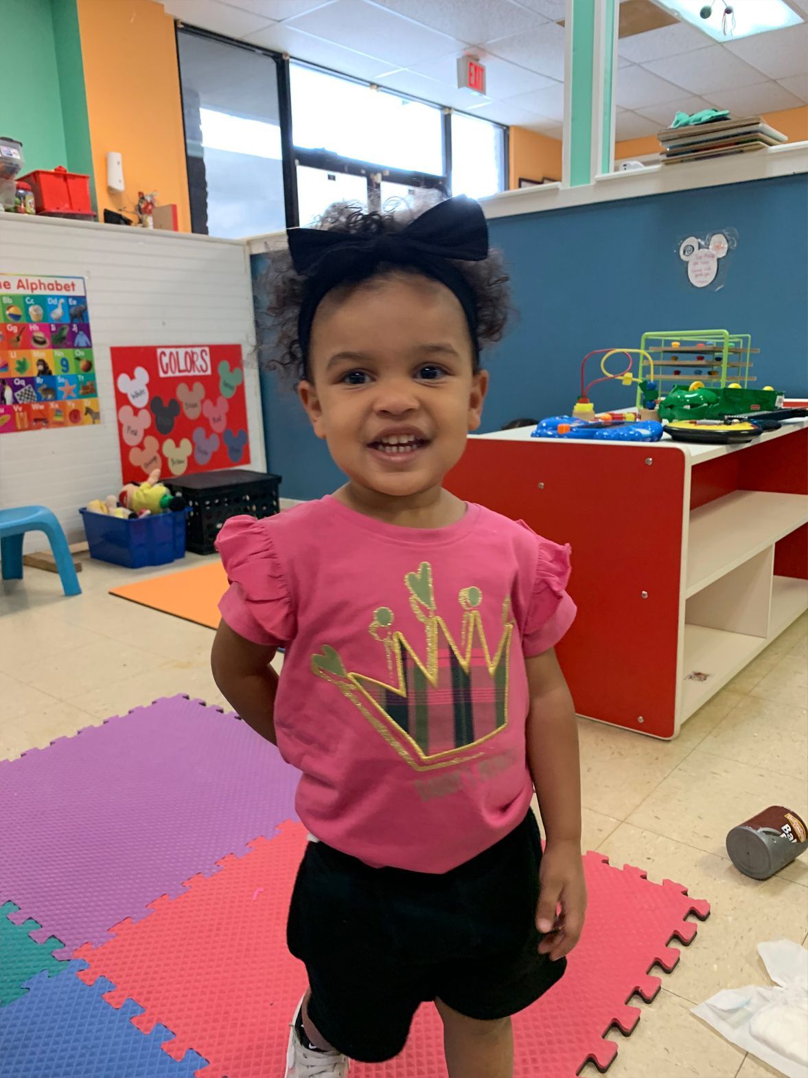 Young child smiles, wearing pink crown shirt, black shorts, and bow. Plays in a colorful playroom.