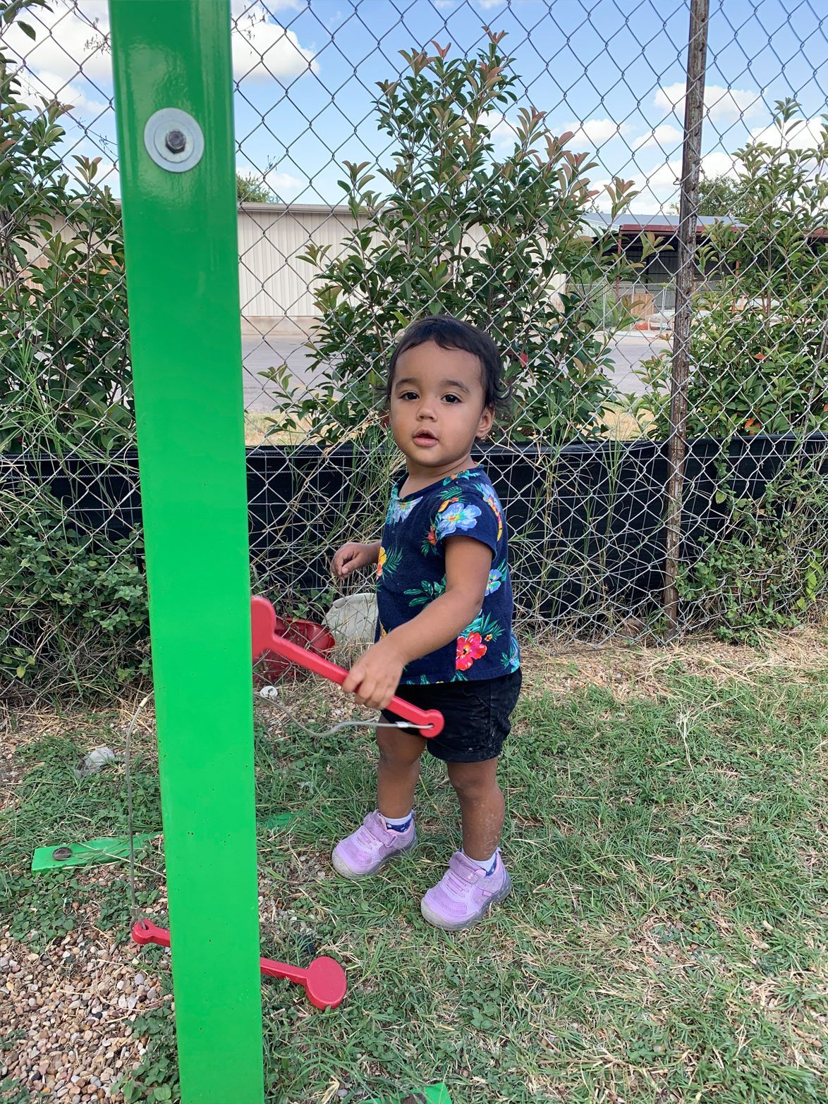 A young child in a floral shirt and shorts stands on grass, holding a red toy mallet near a green post.