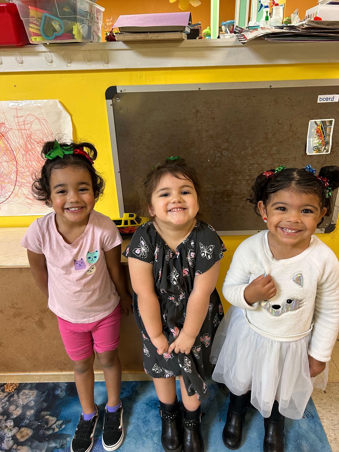 Three smiling children with matching hair accessories stand in front of a chalkboard.