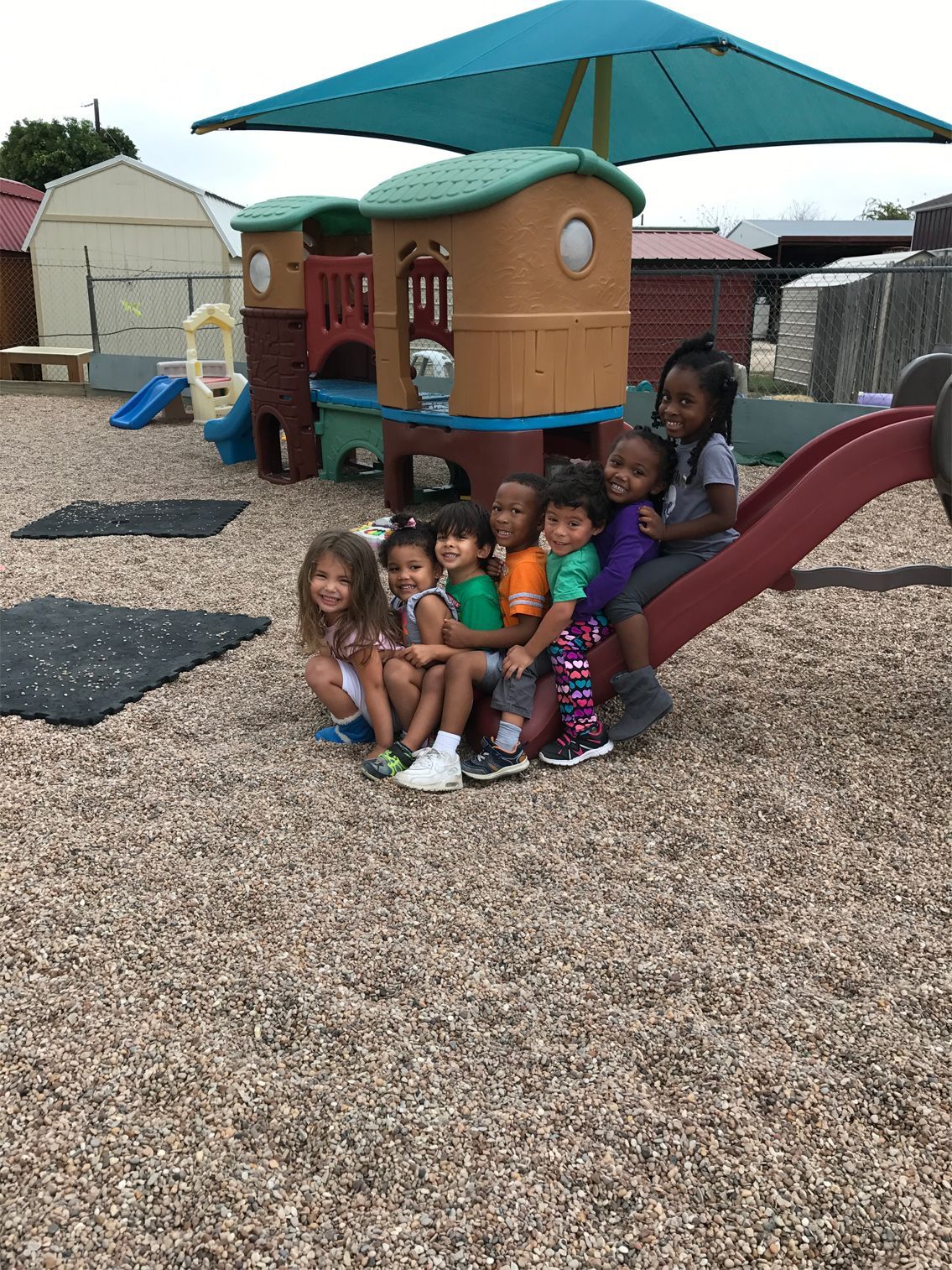 Children smiling, sitting on a playground slide with a playhouse in the background.