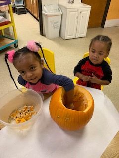 Two children at a table, scooping out a pumpkin. One child smiles, the other watches.