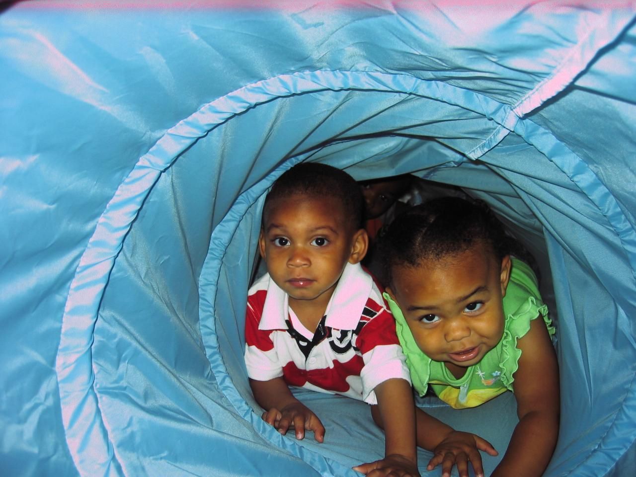 Two children crawling through a blue play tunnel; one in a red shirt, the other in a green shirt.