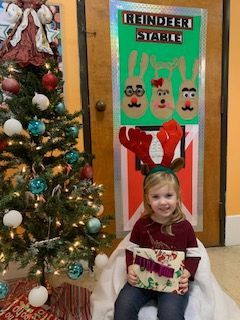 Girl in reindeer antlers sits in front of a Christmas-themed door, next to a decorated tree.