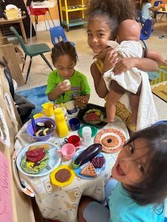 Children playing with toy food at a table. One child holds a doll. Playroom setting.