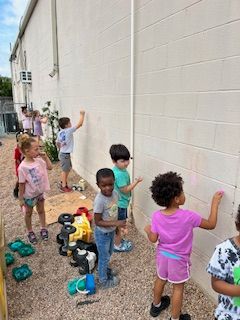 Children drawing with chalk on an outdoor brick wall. Toys are scattered on the ground.