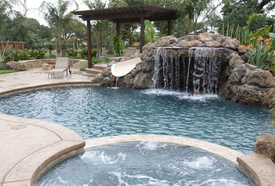 Pool with waterfall, slide, and spa. Rocks surround pool, pergola, and lush landscaping.