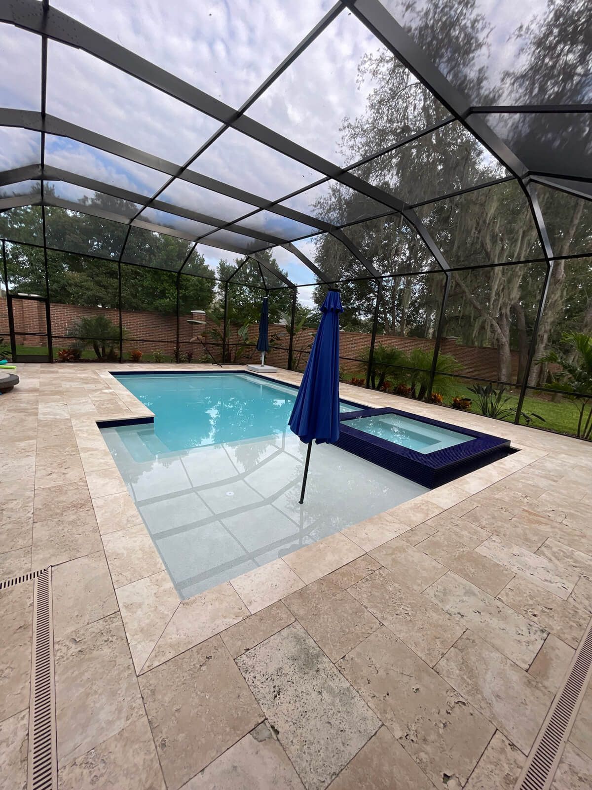 Pool with screen enclosure, blue umbrella, spa, and light stone patio.