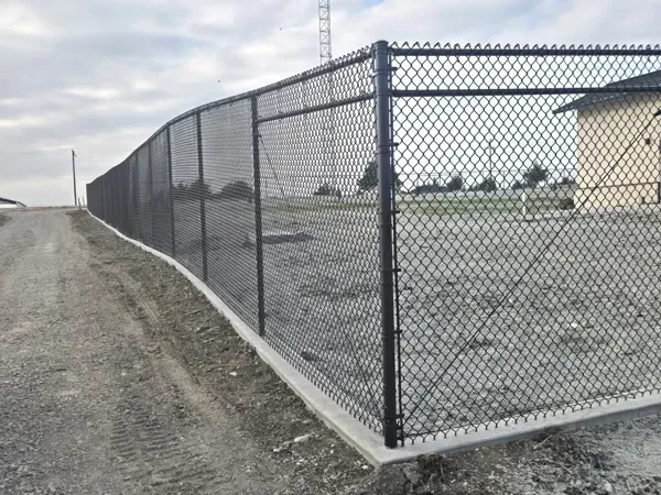Chain-link fence along a gravel road beside an empty paved lot under cloudy skies