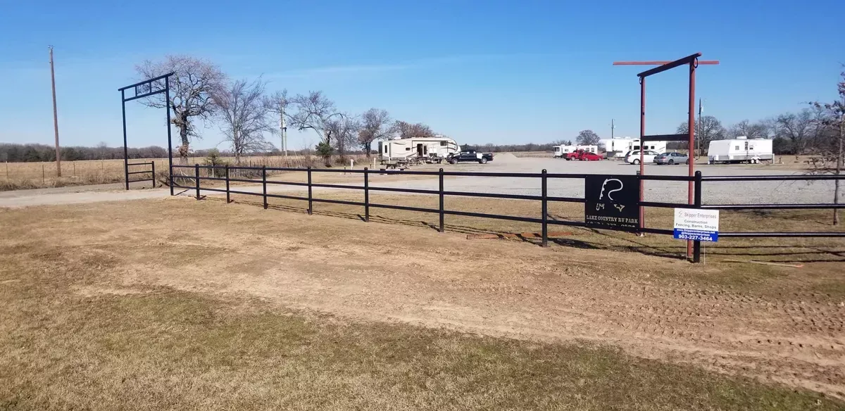 Rural roadside fence and gate beside a grassy field under a clear sky, with houses in the distance