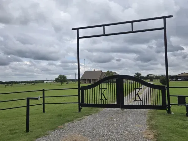 Black metal gate with farm signs at the end of a gravel driveway in a grassy field under cloudy sky