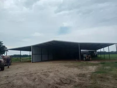 Large open-sided metal shed on a dirt lot under a cloudy sky, with vehicles parked beside it.