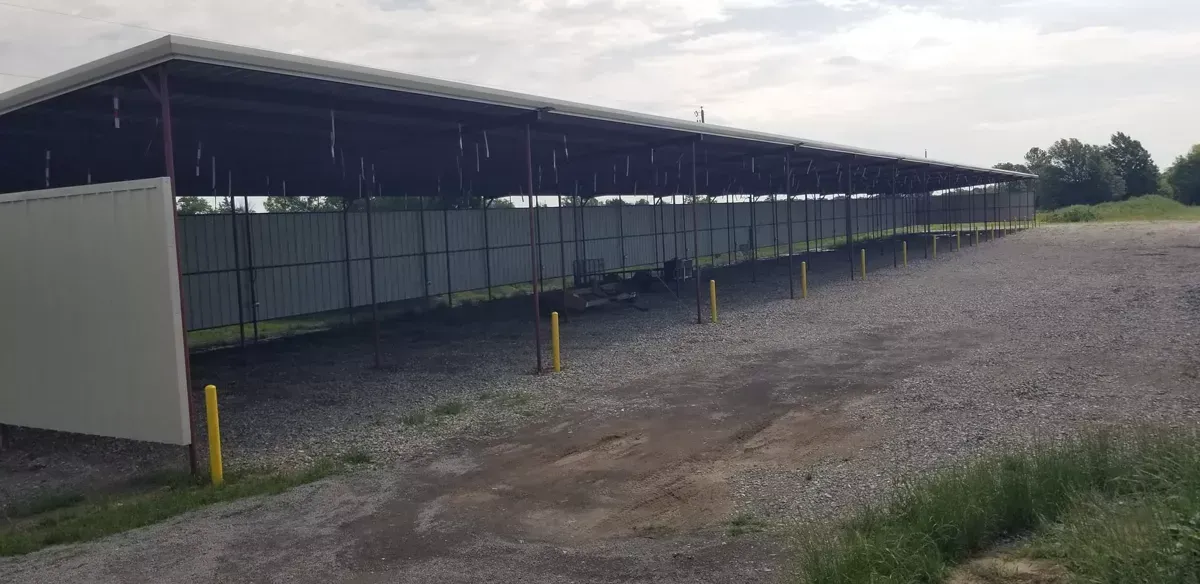 Long, low metal shelter with open sides on a gravel lot under an overcast sky